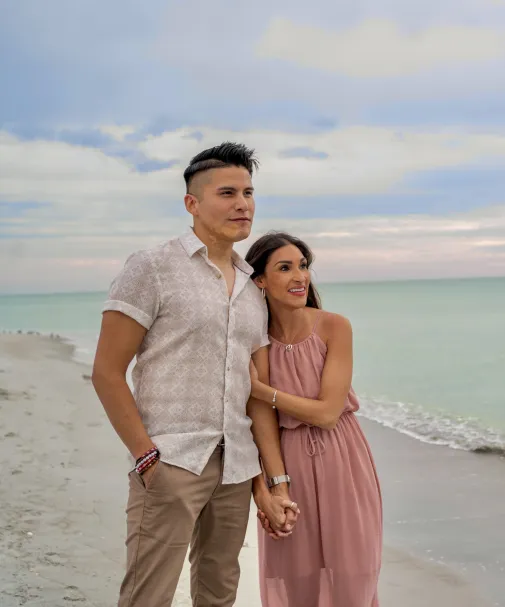 Couple standing hand in hand on a sandy beach at sunset, looking out over calm turquoise water under a cloudy sky.